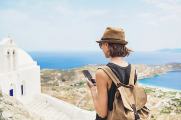 Young tourist woman using smartphone over beautiful landscape with sea in Greece. Traveler girl with backpack and mobile phone. Travel, tourism, summer holidays, active lifestyle concept