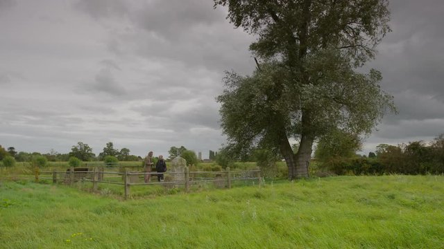 Wide low angle time-lapse still shot of a countryside landscape with a big tree, two men standing at a historical fenced grave stone, and thick dark-grey clouds moving in the sky, Scottish highlands