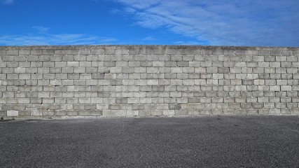 Cinder block wall with an asphalt road in front and blue sky with clouds above it. Background for...