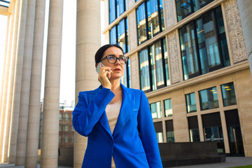 Serious woman in glasses and official wear successful government worker talking via mobile phone while standing outdoors near enterprise during work break. Female lawyer having cellphone conversation