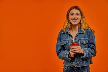 A woman with wavy fair hair in a denim jacket smiles at the camera with a red cardboard cup of coffee in her hands.