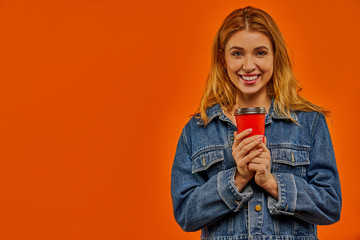 A woman with wavy fair hair in a denim jacket smiles at the camera with a red cardboard cup of coffee in her hands.