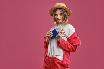 Blonde female in straw hat, white blouse and red pantsuit. She is holding passport and ticket while posing against pink studio background. Close-up