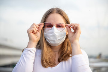 Portrait of a beautiful caucasian girl in a mask and pink glasses in the shape of hearts looks at the camera. in the background planes and the airport in blur. koronaviirus сovid-19 concept