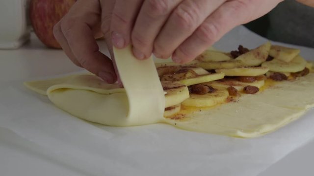 senior woman hands make delicious strudel with apples from rolled dough on table with parchment in kitchen, side view, close up