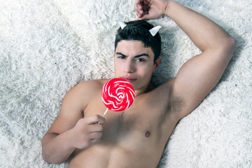 Portrait of handsome muscular young devil with horns lying on a shaggy blanket licking a lollipop