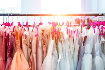 Rack with many beautiful holiday dresses apparel on hangers at female fashion showroom indoor. Kid girl dress hire studio for celebration birthday party or photography session event. Bright sun day