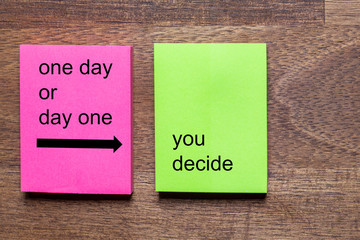 pink and yellow self-adhesive note sticker with the motto: one day or day one you decide. Background dark wooden table