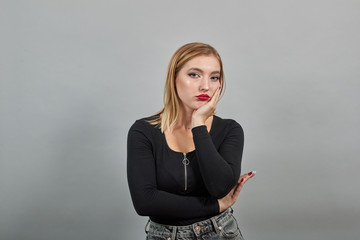Young blonde girl in black jacket on grey background sad woman is bored, touches chin with hand