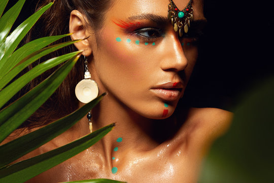 Closeup portrait of woman tanned skin colored makeup tropical jungle paint on the face sweat plant palm leaves wild style exotic accessory beautiful sexy model ethnic model.
