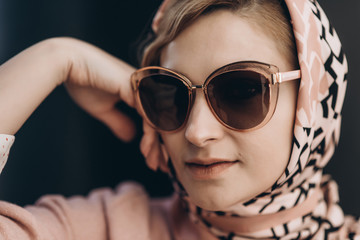 Elegant fashion. Stylish blonde woman in pink clothes, scarf and sunglasses posing in the city against a background of black huge barrels. Soft selective focus. Close-up.