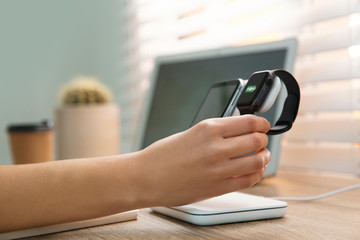 Obraz premium Woman putting smartwatch onto wireless charger at wooden table, closeup. Modern workplace accessory