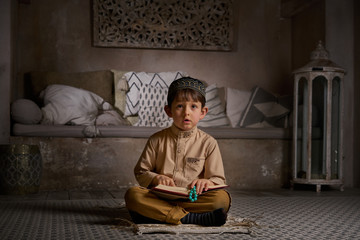 Fototapeta na wymiar Boy in arabic clothes with rosary beads reading holy quran book praying to Allah, prophet Muhammad holy spirit religion symbol concept inside eastern traditional interior