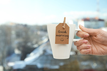 Woman holding cup of delicious coffee with GOOD MORNING wish near window