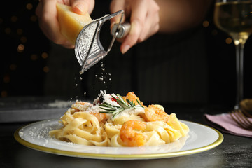 Woman grating cheese onto pasta with shrimps on black table, closeup - obrazy, fototapety, plakaty