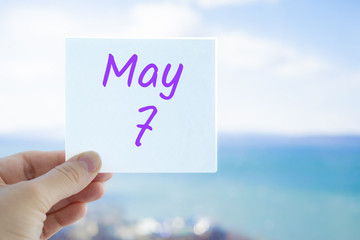 May 7th. Hand holding sticker with text May 7 on the blurred background of the sea and sky. Copy space for text. Month in calendar concept