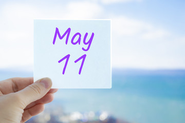 May 11th. Hand holding sticker with text May 11 on the blurred background of the sea and sky. Copy space for text. Month in calendar concept