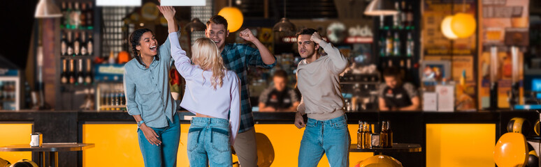 panoramic shot of happy multicultural friends showing winner gestures in bowling club