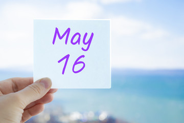 May 16th. Hand holding sticker with text May 16 on the blurred background of the sea and sky. Copy space for text. Month in calendar concept