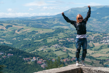 Girl on the edge of the top of the world standing with raised hands. Destination scene. incredible view from the mountain. Motorcycle travel concept. Vacation in Italy. European tour to San Marino