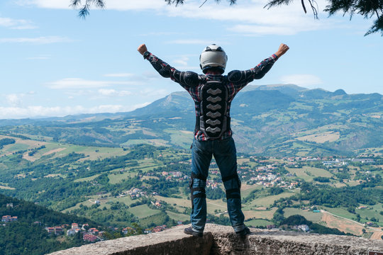 Man on the edge of the top of the world standing with raised hands. Destination scene. incredible view from the mountain. Motorcycle travel concept. Vacation in Italy. European tour to San Marino