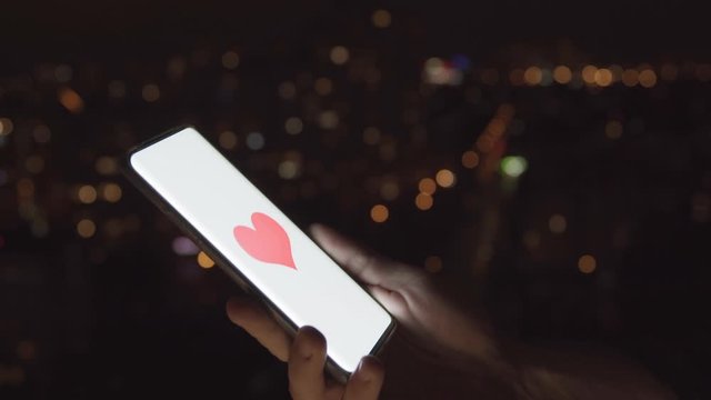 Close-up of Smartphone at Night with Red Heart Appearing on White Screen. Man Picks up his Phone to Find a Date. Concept of Online Mating