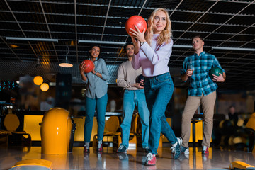 smiling blonde girl throwing bowling ball near multicultural friends