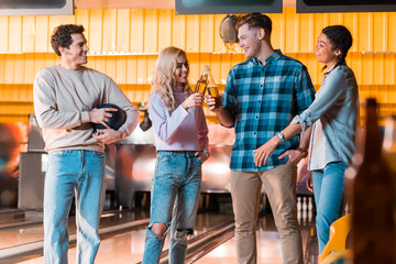 happy multicultural friend talking and clinking bottles of beer in bowing club