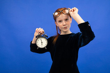 serious red-haired teenager girl in glasses in a black dress recalls punctuality holding an alarm...