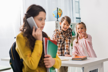 selective focus of cruel classmate pointing with finger at upset schoolgirl talking on smartphone,...