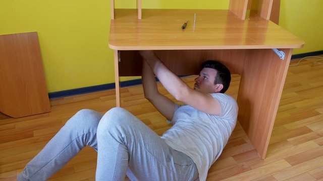 Man collecting parts of computer table screwing with bolts lying under table. Repairman is assembling furniture himself and making renovation at home.