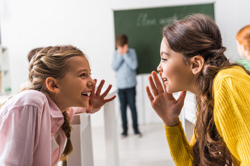 selective focus of schoolgirls gossiping near upset classmate, bullying concept