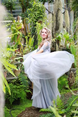  beautiful blonde long hair girl in a flying tulle skirt walks in a botanical garden among green plants