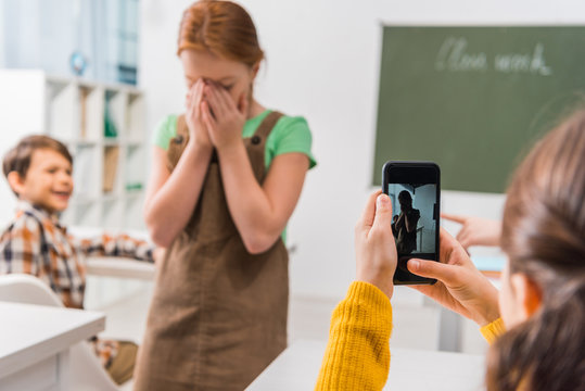 selective focus of schoolgirl taking photo of classmate, cyberbullying concept