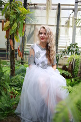 Obraz na płótnie Canvas beautiful girl, blond long hair in a gray top and a tulle aerial skirt sits in a botanical garden among green plants