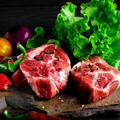 Two juicy steaks lie on a wild stone next to red and green chili peppers and a fresh green salad on a black wooden table