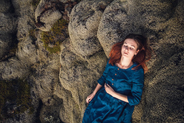 Young beautiful woman in blue dress lying on the volcanic formations covered by green moss on a background green. Place for text or advertising