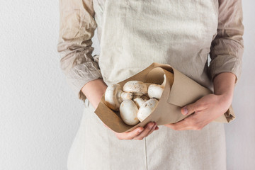 Young Caucasian woman in linen apron holds in hands brown paper bag with freshly picked white button mushrooms in kitchen. Kinfolk style. Meal cooking healthy plant based diet concept