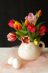 Spring holiday mood. Multicolored tulips in a jug with two whole raw white eggs on a black-white background. Preparing for Easter. Flower still life, vertical orientation