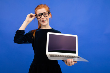 smart red-haired teenager girl in glasses holds a laptop with a blank screen mock up to insert a page on a blue background