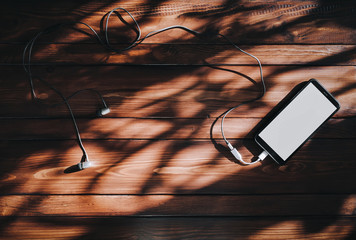 Black smartphone with white screen and dark headphone lies on a brown wooden background mahogany...