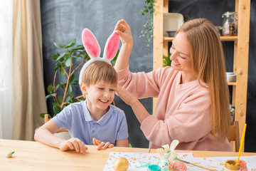 Happy young mother adjusting rabbit ears headband on son head while they dressing for Easter party