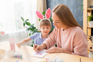 Young creative mother sitting with curious son in rabbit ear headband at table and signing Easter card with paintbrush