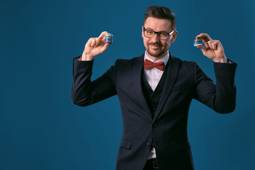 Man in black classic suit, red bow-tie, glases is showing some colored chips, posing on blue studio background. Gambling, poker, casino. Close-up.
