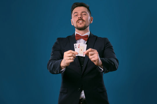 Man in black classic suit and red bow-tie showing two playing cards while posing against blue studio background. Gambling, poker, casino. Close-up.
