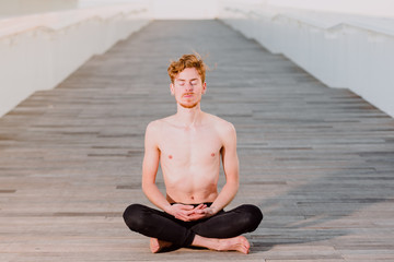 Fototapeta na wymiar Young red-haired man practicing the lotus yoga posture, Padmasana, outdoors on the wooden floor