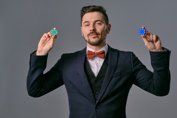 Fototapeta premium Man in black classic suit and red bow-tie showing some colored chips, posing against gray studio background. Gambling, poker, casino. Close-up.