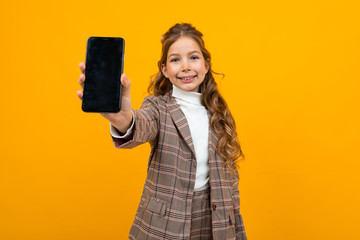 cute smiling young girl in a classic brown jacket shows a phone with a mockup on a yellow background with copy space