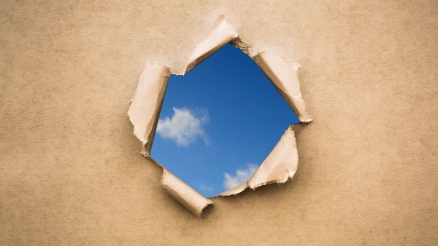 Creative 4k time laps video of fast moving clouds in the blue sky that are visible through a hole with torn edges in old retro grunge vintage paper.