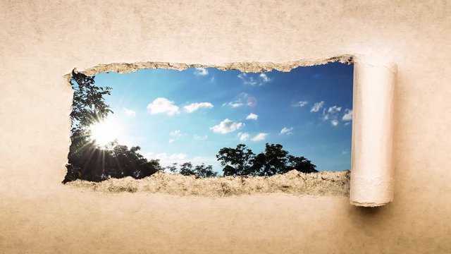 Creative 4k time laps video of fast moving clouds in the blue sky at sunset witn sunflare that are visible through a hole with torn edges in old retro grunge vintage paper.